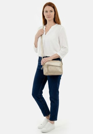 Frau mit roten Haaren, trägt eine weiße Bluse, dunkle Jeans und weiße Sneaker, hält eine goldene Umhängetasche mit langem Riemen vor weißem Hintergrund.