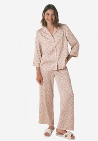 Pyžamový set ze světle růžové látky s drobnými květinovými vzory. Dlouhý rukáv s knoflíky a volné kalhoty, doplněné bílými pantoflemi.