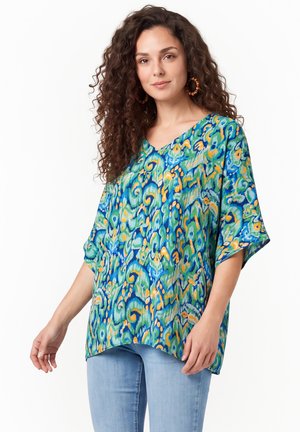 Kvinde med langt krøllet hår iført en løs bluse med blå, grønne og gule mønstre og lyseblå jeans, stående mod en hvid baggrund.