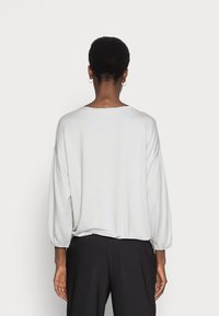 Light gray, long-sleeve top with a loose fit, rounded hem, and elasticized cuffs, paired with black pants. Back view of the garment.