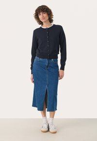 Mornarsko modri kardigan s gumbi, v kombinaciji z modrim denim krilom z razporkom spredaj. Bele nogavice in superge dopolnjujejo outfit.