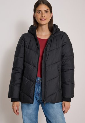 Femme portant une veste matelassée noire à capuche par-dessus une chemise rouge et un jean bleu, debout devant un fond clair uni.