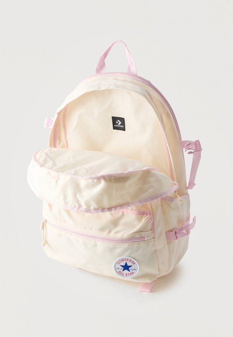Zaino Converse crema e rosa con scomparto principale aperto, tasca anteriore con cerniera e patch con logo su sfondo bianco.