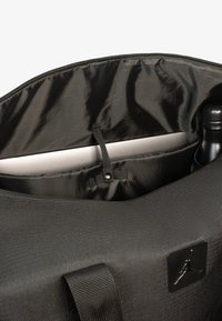 Bolsa textil negra con una forma estructurada, que cuenta con una abertura con cremallera, un compartimento interior para un portátil y un bolsillo lateral para una botella.