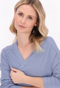 Jasnoniebieska, prążkowana bluzka z długim rękawem i dekoltem w kształcie litery V, z teksturowanym wykończeniem i luźnym krojem. Ręka modelki spoczywa na jej ramieniu.