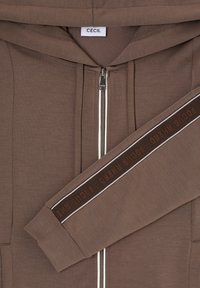 Brązowa bluza z kapturem i białym zamkiem błyskawicznym, z kapturem oraz rękawem ozdobionym ciemnobrązowym paskiem z napisem "POCUS AHEAD". Gładka tekstura.