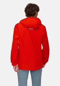 Czerwona wodoodporna kurtka z kapturem, z logo na plecach, dopasowanymi mankietami i prostym krojem. Zestawiona z jasnoniebieskimi spodniami.