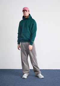 Green hoodie with kangaroo pocket and ribbed cuffs, paired with gray corduroy pants. The individual wears a light pink cap and silver sneakers.