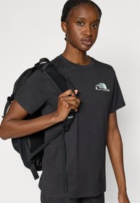 Μαύρο βαμβακερό μπλουζάκι με κοντά μανίκια και στρογγυλή λαιμόκοψη, με μικρό λευκό και τιρκουάζ λογότυπο στο αριστερό μέρος του στήθους.