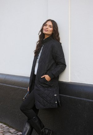 Quilted black long coat with a high collar and zip closure, featuring side pockets and a curved hem. Worn over a light sweater and black pants.