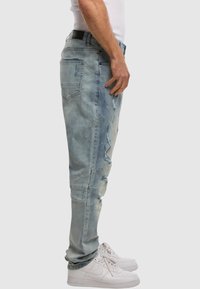 Vaqueros de denim desgastado en un tono azul claro, con un corte ajustado, costuras visibles y múltiples bolsillos. Combinados con zapatillas blancas.