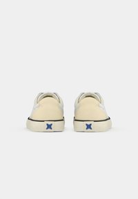 Une paire de baskets beige et blanche vue de l'arrière, présentant des logos en forme d'étoile bleue sur la semelle près du talon.