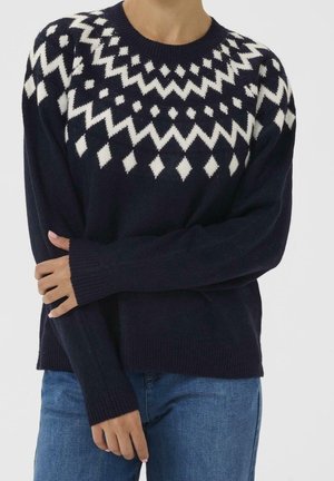 Granatowy sweter z białym geometrycznym wzorem diamentu i zygzaka na klatce piersiowej i ramionach, noszony z niebieskimi dżinsami.