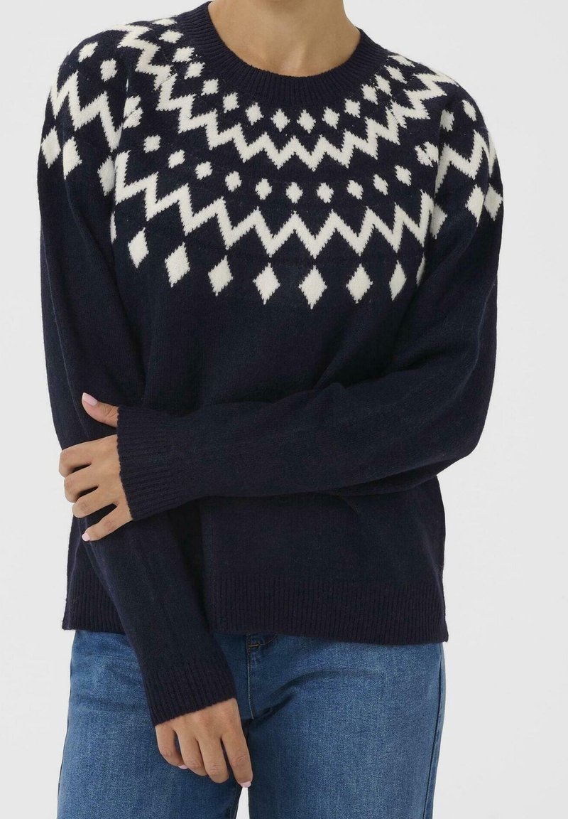 Pull bleu marine avec motif géométrique blanc en forme de losange et zigzag sur la poitrine et les épaules, porté avec un jean bleu.