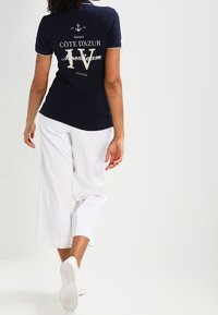 Polo shirt bleu marine avec texte et graphismes blancs, associé à un pantalon large blanc et des baskets blanches, vu de dos.