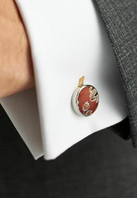 Bouton de manchette à motif rouge avec un cadre circulaire en argent, monté sur le poignet d'une chemise blanche, contrastant avec un costume gris.