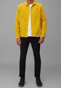 Chaqueta camisa de pana amarilla con dos bolsillos en el pecho, combinada con pantalones ajustados negros y zapatillas negras con suela marrón.