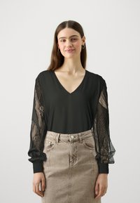 Svart topp med V-hals, med gjennomsiktig blonder-ballongsleeves og tettsittende mansjetter. Kombinert med et lysebrunt denimskjørt.