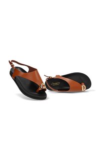 Sandalias de cuero marrón de tiras con suelas negras y acentos de hebilla dorada en la correa del dedo, colocadas una al lado de la otra sobre un fondo blanco.