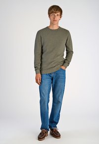 Oliwkowy sweter z dzianiny z okrągłym dekoltem, zestawiony z jasnoniebieskimi dżinsami i brązowymi butami żeglarskimi. Model stoi swobodnie z ręką w kieszeni.