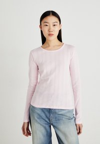 Chemise rose clair à manches longues avec une texture côtelée et de subtils motifs verticaux, col rond, associée à un jean bleu clair.