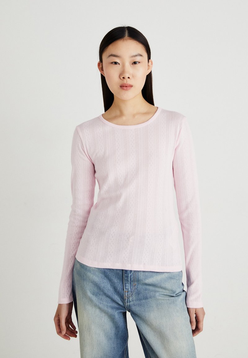 Chemise rose clair à manches longues avec une texture côtelée et de subtils motifs verticaux, col rond, associée à un jean bleu clair.