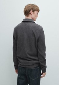 Tmavě šedý pletený svetr s límečkem, dlouhými rukávy a volným střihem, kombinovaný s tmavými džínami. Textura působí měkce a útulně.