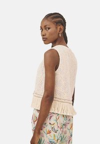 Chaleco beige de punto con patrón floral, tejido texturizado y dobladillo con flecos. La modelo lleva pantalones con estampado floral, mostrando un look en capas.