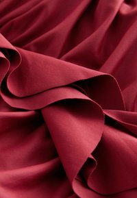Vīna krāsas audums ar gludu tekstūru, raksturojot mīkstas drapērijas un locījumus, attēlojot bagātīgu, vienkrāsainu toņu bez rakstiem vai rotājumiem.