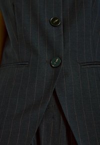 Temno modra tkanina s pinstripes vzorom, ki jo krasita dva sijoča črna gumba, kroj z ostro obrobo in ozkimi žepi. Vidni akcenti šivanja.