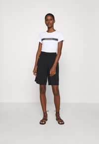 T-shirt bianco con logo "Calvin Klein Jeans" nero, abbinato a pantaloni corti neri fino al ginocchio. Il modello indossa sandali neri con cinturino. Sfondo bianco.