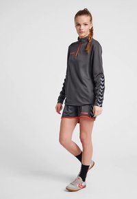 Szary sportowy top z półzamkiem i pomarańczowymi akcentami, w komplecie z dopasowanymi szortami. Charakteryzuje się teksturowaną tkaniną oraz skośnym wzorem pasków na rękawach.