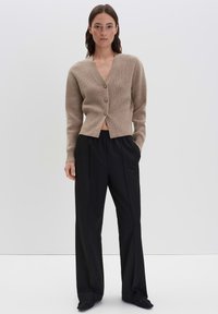 Femme se tenant contre un fond blanc, portant un cardigan beige à boutons, un pantalon noir à jambe large et des chaussures noires, les mains dans les poches.