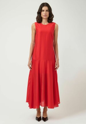 Woman standing wearing a sleeveless, flowing red maxi dress and black pointed-toe heels against a plain light background.
