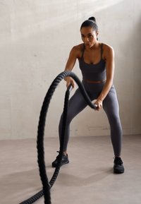Black battle ropes are held in both hands by a person in charcoal-grey athletic wear, performing an exercise on a light grey floor.