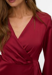 Rød satinvendekjole med lange ærmer, dyb V-udskæring og en tætsiddende talje. Stoffet har en glat, blank finish.