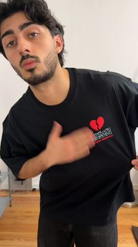 Ung mann med mørkt hår og skjegg trekker i en svart T-skjorte med et rødt, knust hjerte og japansk tekst nær brystet, innendørs med tregulv.