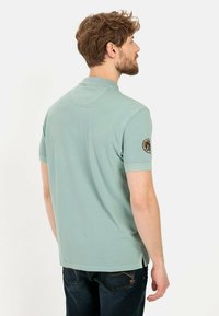Lichtgroen polo-shirt met korte mouwen en een kraag, voorzien van een textuurafwerking. Het shirt heeft een badge op de linker mouw en zijsplitten aan de onderkant.