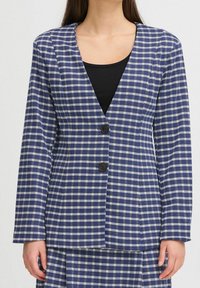 Femme portant un blazer à carreaux bleu et blanc avec deux boutons foncés sur un haut noir, visible du cou aux hanches.