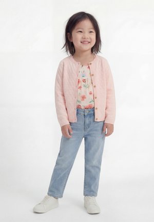 Χαμογελαστή κοπέλα φοράει ροζ καπιτονέ ζακέτα, φλοράλ μπλούζα, ανοιχτό μπλε τζιν και λευκά αθλητικά παπούτσια, στέκεται μπροστά σε λευκό φόντο.