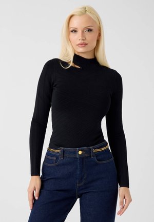 Jonge vrouw met lang blond haar draagt een zwart langemouwenshirt en blauwe jeans met een gouden kettingriem, staand tegen een lichtgrijze achtergrond.