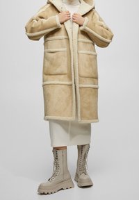 Beige faux shearling jakke med hætte, der har en struktureret overflade, fire lommer og kontrastkant. Parret med beige kampstøvler.
