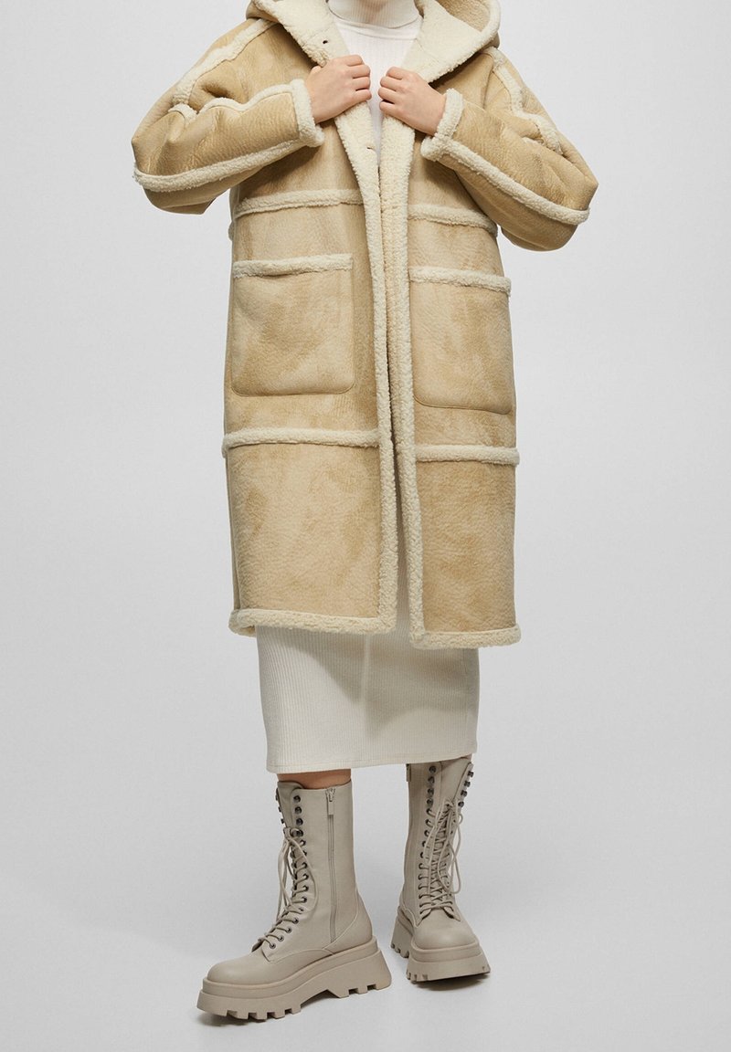 Beige faux shearling jakke med hætte, der har en struktureret overflade, fire lommer og kontrastkant. Parret med beige kampstøvler.