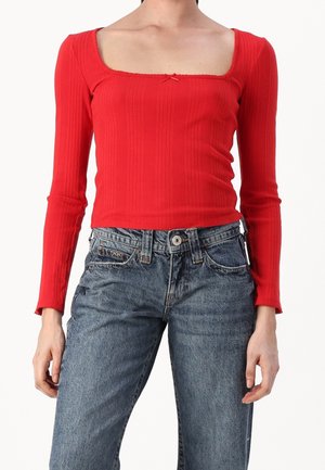 Kobieta ubrana w dopasowany czerwony prążkowany top z długim rękawem i kwadratowym dekoltem oraz niebieskie wysokowalne dżinsy, stojąca z luźno opuszczonymi ramionami.