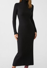 Robe noire en tricot côtelé avec un col haut, des manches longues et une coupe ajustée qui descend en dessous du genou. Le tissu texturé met en valeur la silhouette.