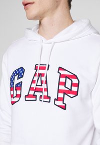 Sweat à capuche blanc arborant un grand logo "GAP", avec des rayures rouges et blanches et un motif d'étoiles bleues sur la lettre « G ». Détail de capuche avec cordon de serrage.