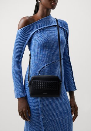 Black leather crossbody bag with woven texture, rectangular shape, and adjustable strap, worn with a blue ribbed long-sleeve dress.