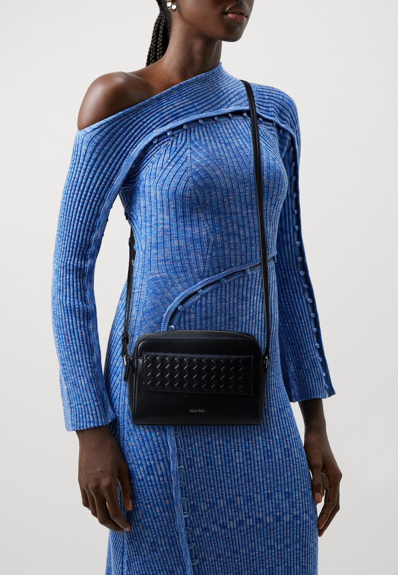 Borsa a tracolla in pelle nera con texture intrecciata, forma rettangolare e tracolla regolabile, indossata con un vestito blu a maniche lunghe a coste.