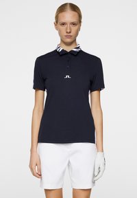 Mörkblå polo T-shirt med randig krage och vita detaljer, utrustad med en treknappsknäppning och en logotyp i mitten av bröstet. Kombinerad med vita shorts och en handske.
