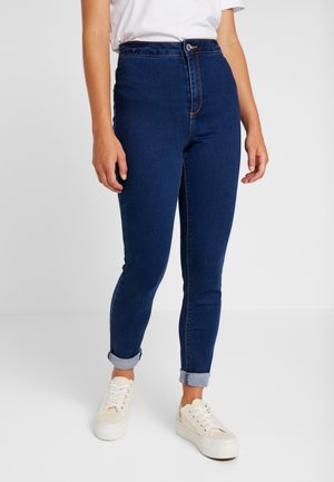 Personne portant un jean skinny bleu foncé avec revers, des baskets blanches et une chemise blanche à manches courtes, debout sur un fond uni.